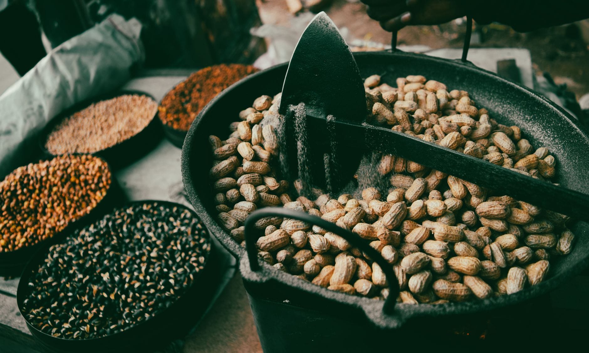 Peanuts in their shells being roasted in a large black pan beside bowls of other seeds and grains
