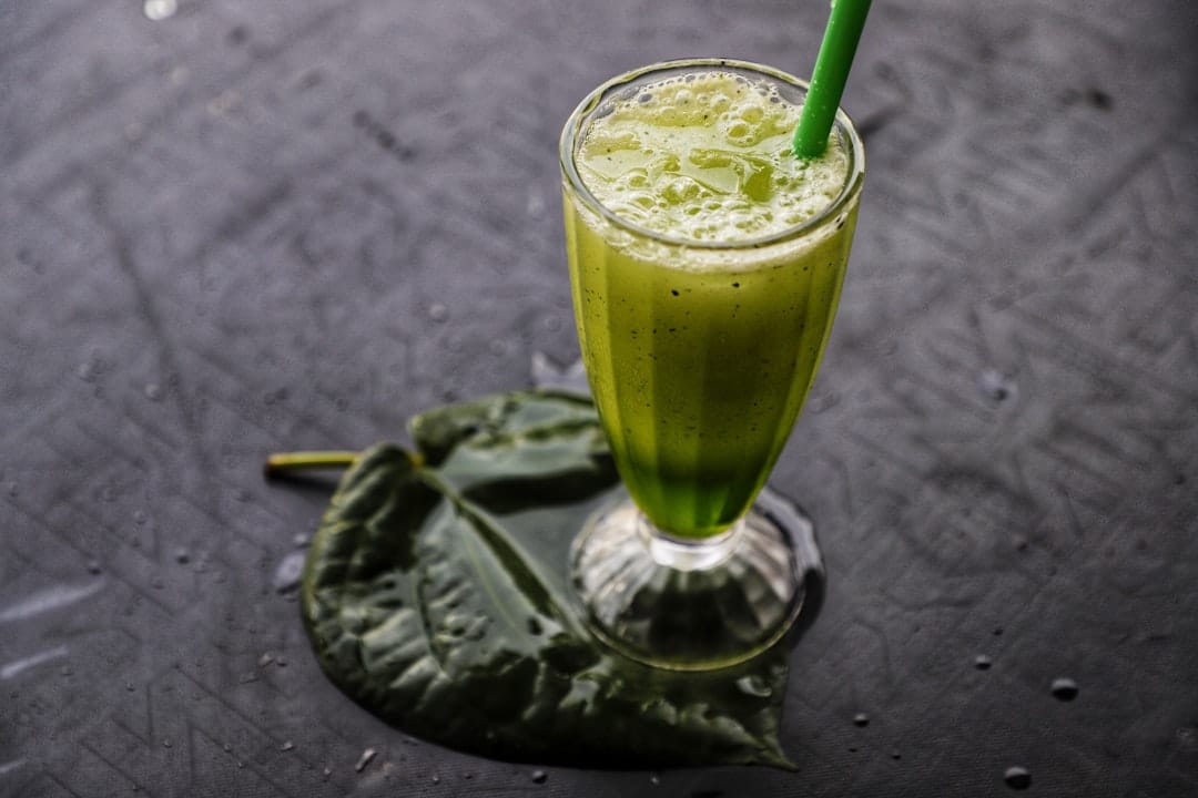 Glass of green juice with a straw resting on a dark leaf