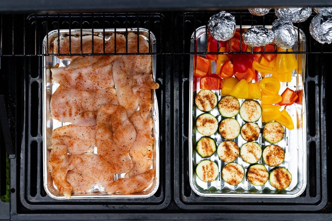 Raw chicken and vegetables cooking in foil trays on a grill