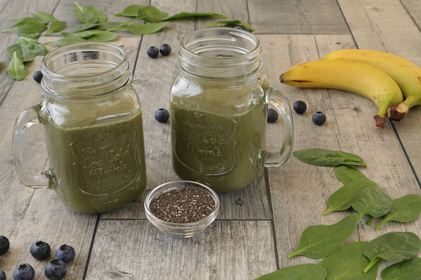 Two glass mugs of green smoothie with spinach leaves, blueberries, bananas and a small bowl of chia seeds