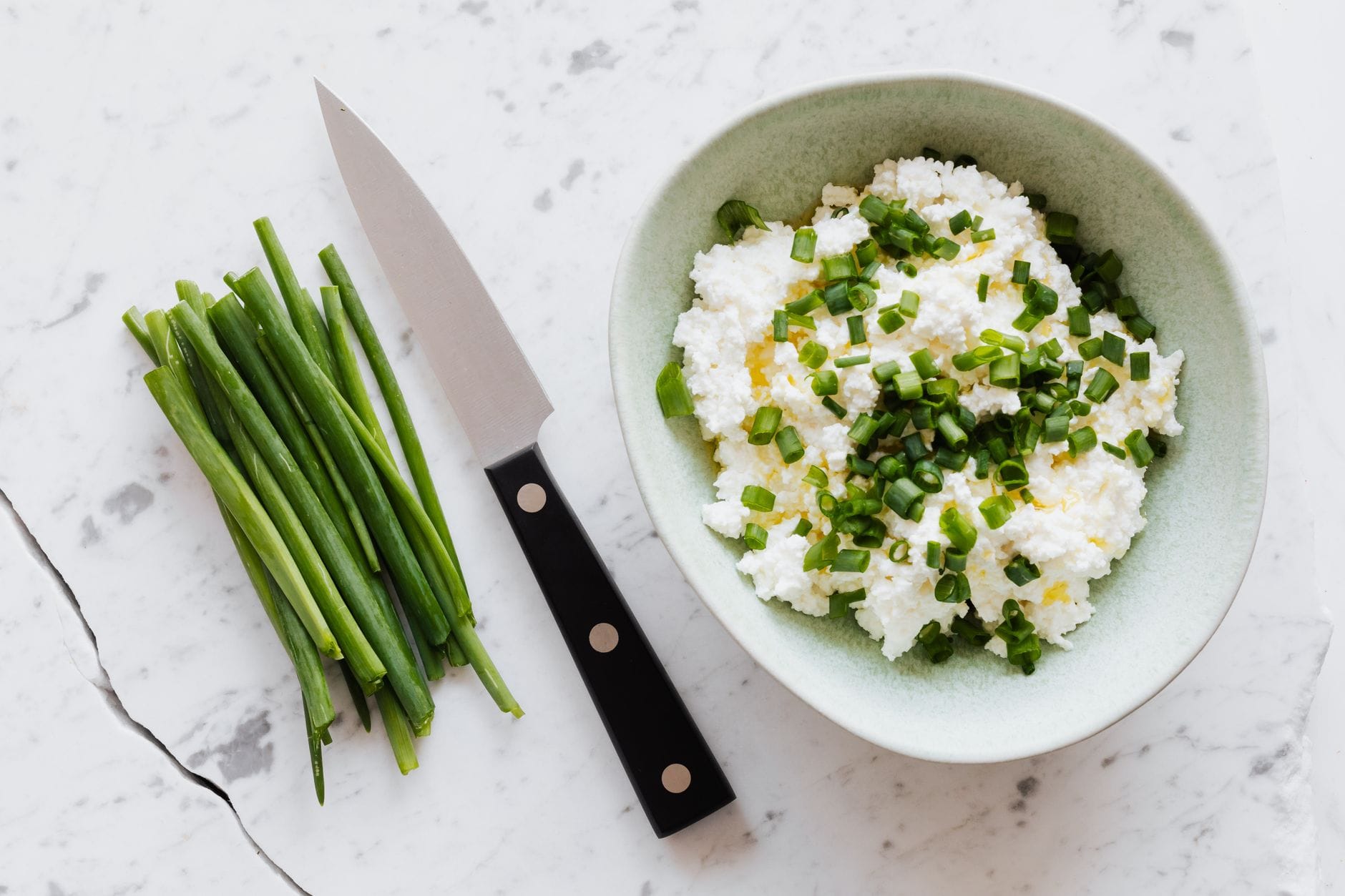 Green bowl of crumbly cottage cheese topped with chopped spring onion beside a knife