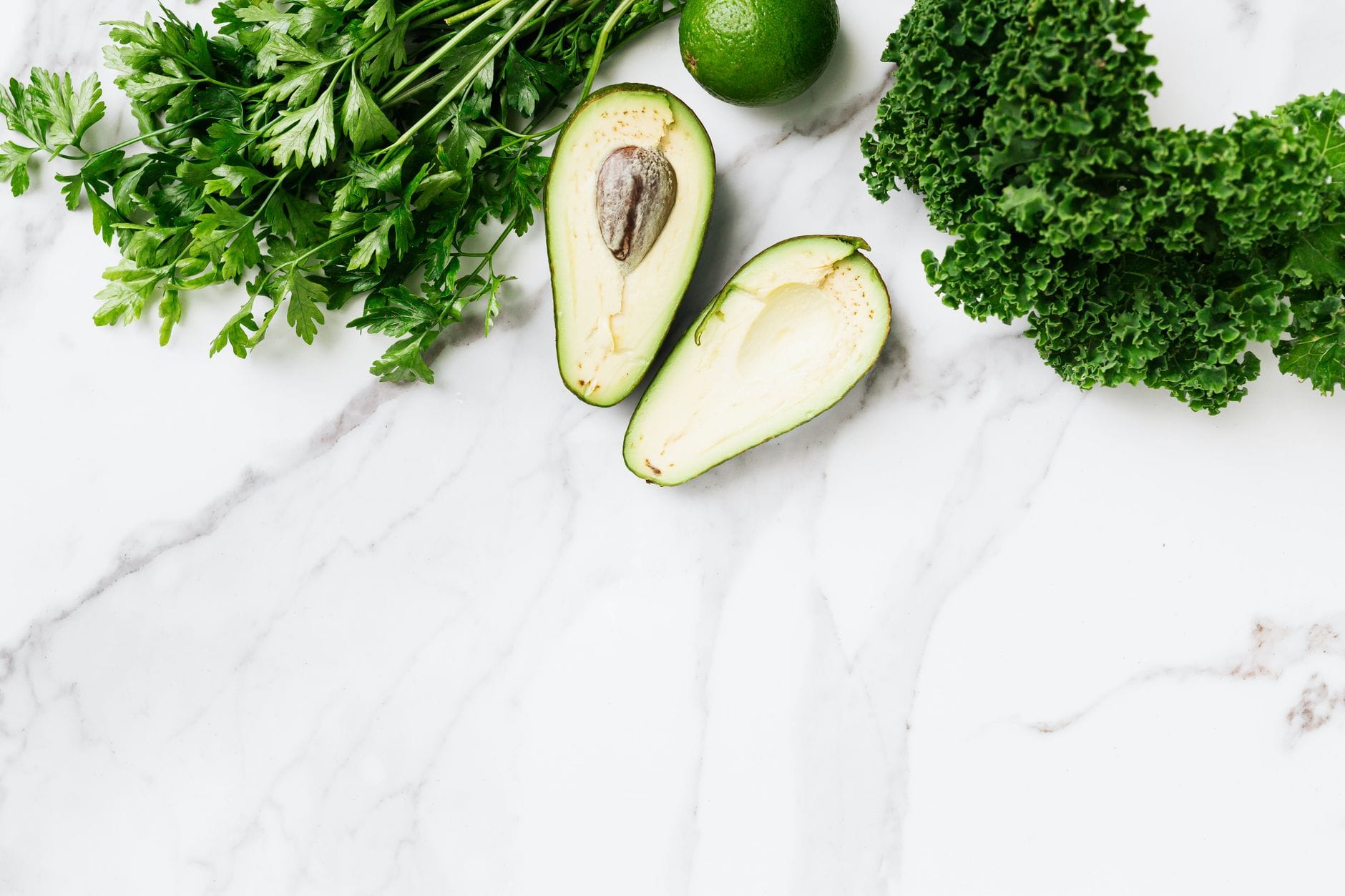 Halved avocado, lime, curly kale and parsley arranged on white marble