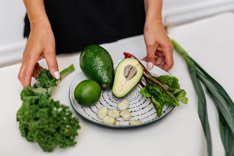 Hands arranging avocado, lime, chard and garlic on a plate beside kale and leeks