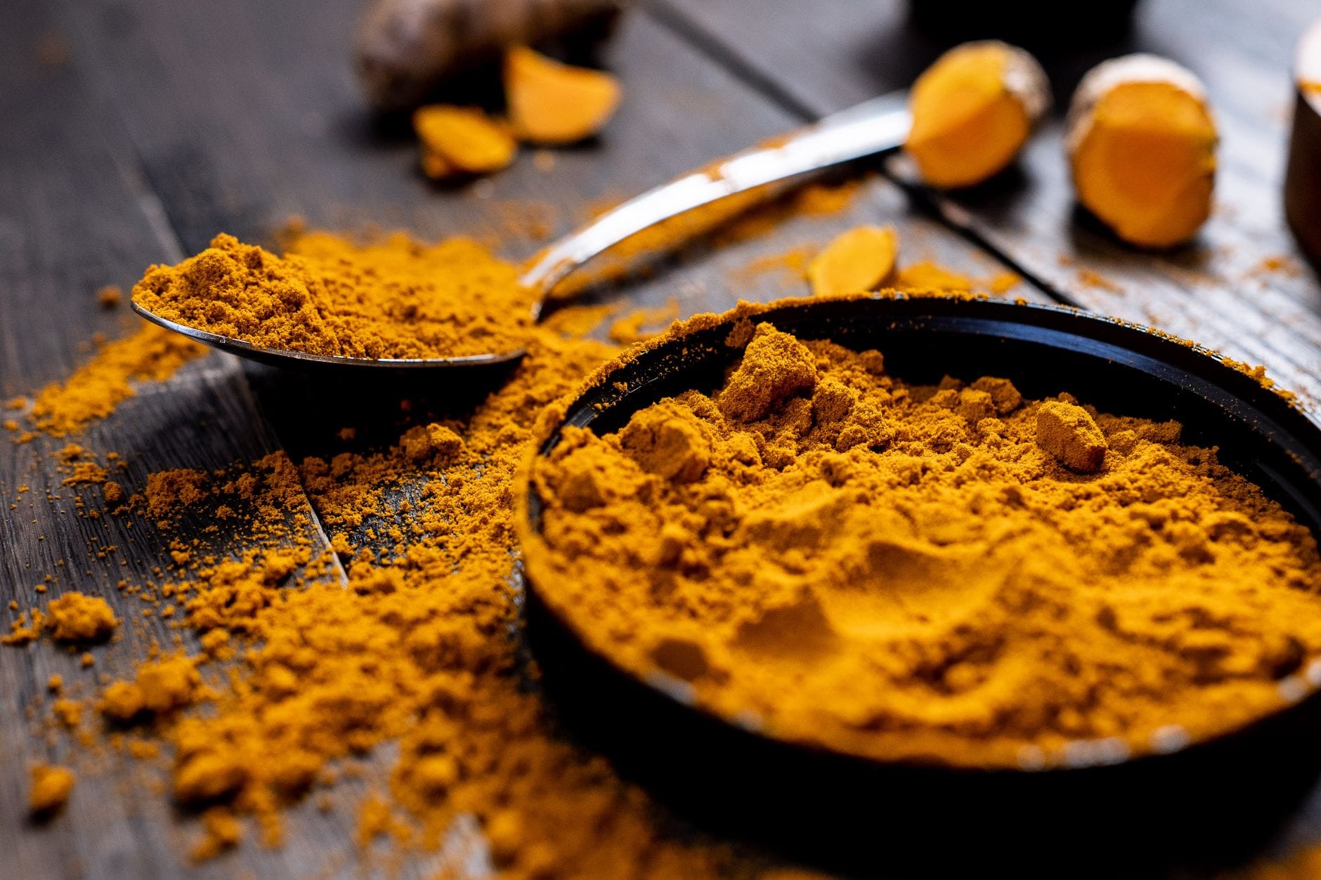 Bowl and spoon of ground turmeric with fresh turmeric root on dark wood
