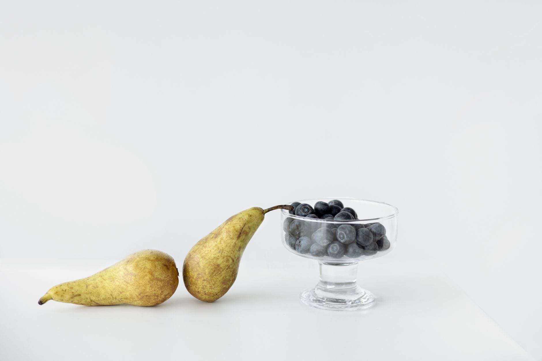 Two pears beside a glass dish of blueberries on a white surface