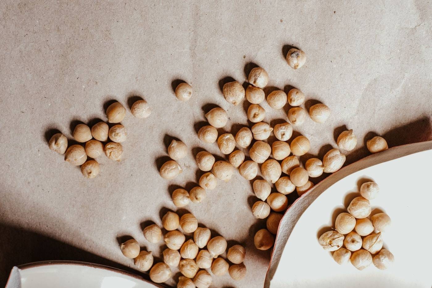 Dried chickpeas scattered across beige paper beside two white ceramic bowls
