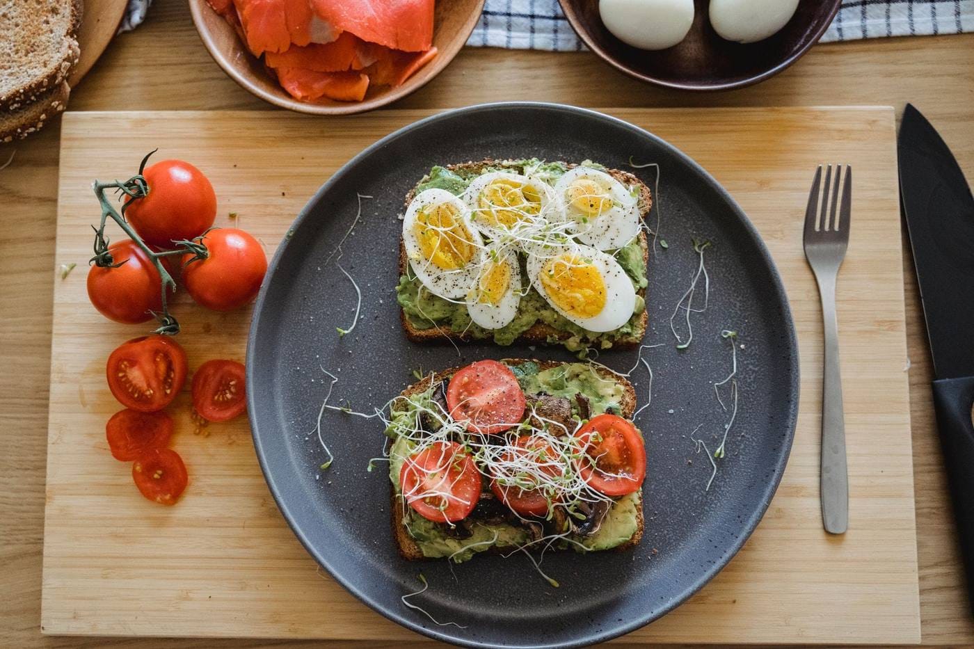 Two slices of avocado toast, one topped with sliced boiled eggs and sprouts, the other with tomato and mushrooms