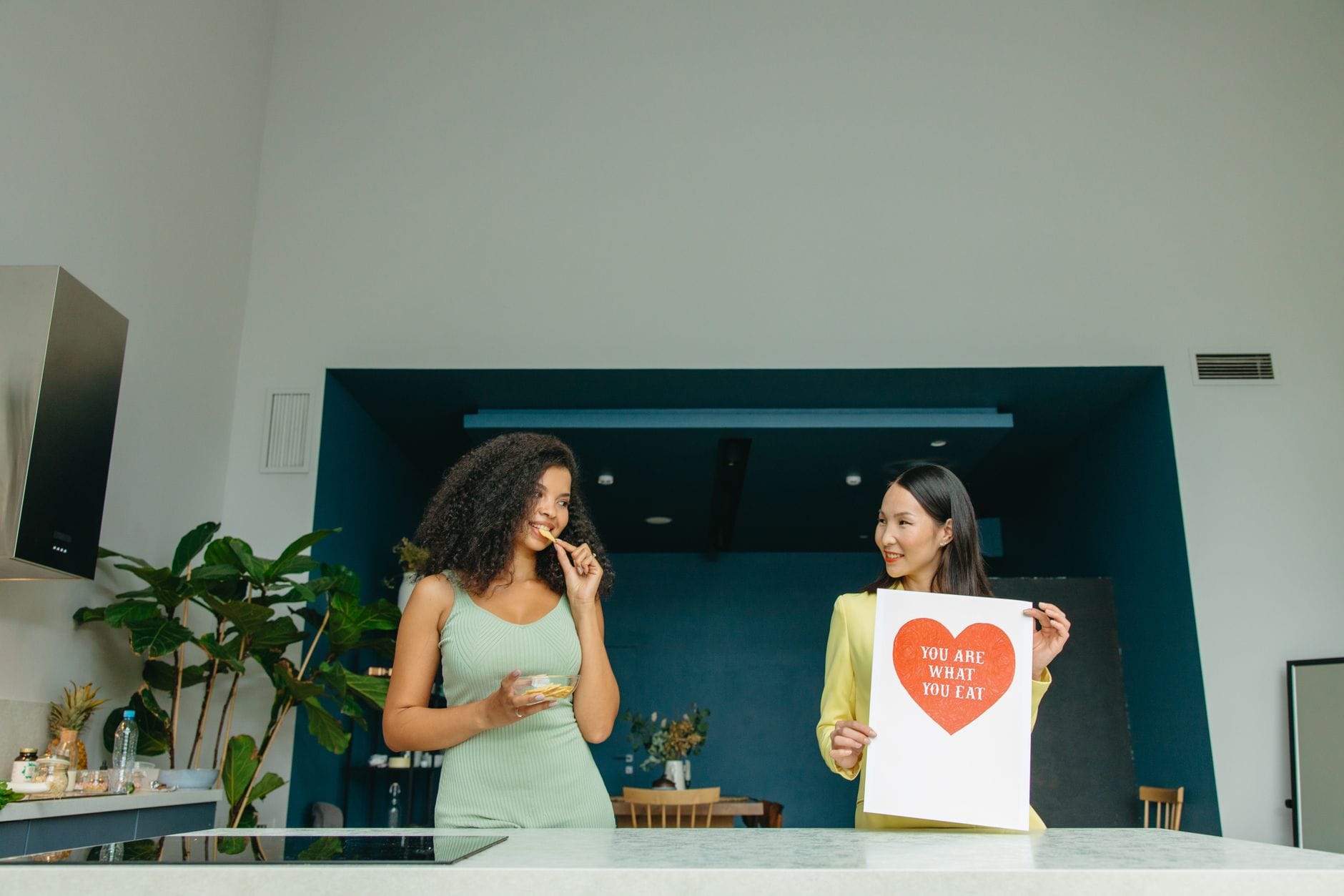 Woman in yellow holds a red heart poster while a friend snacks nearby