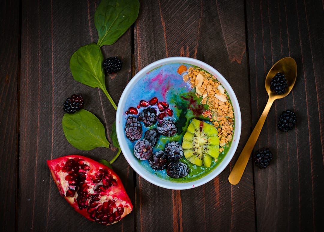 Smoothie bowl with blue-green swirls topped with blackberries, kiwi and granola, with spinach leaves and a gold spoon on dark wood