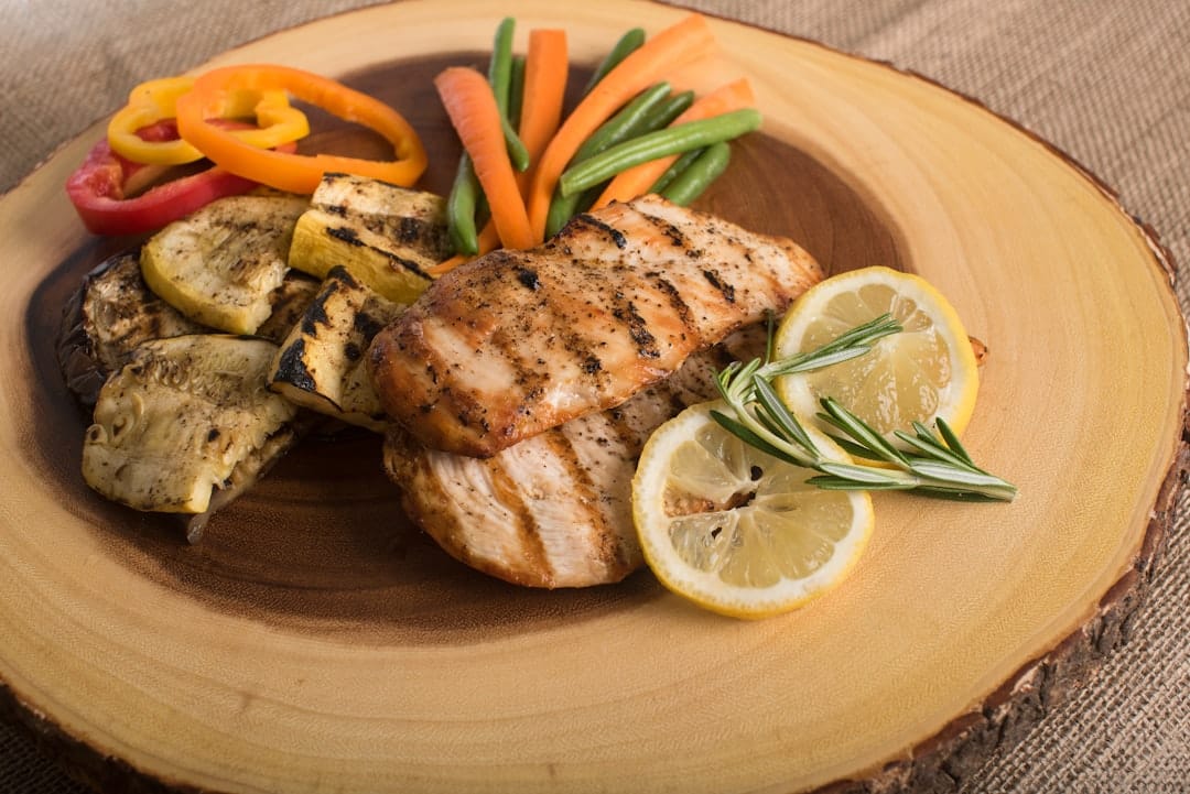 Grilled chicken breast with grilled courgette, peppers, carrots, green beans and lemon on a wooden board