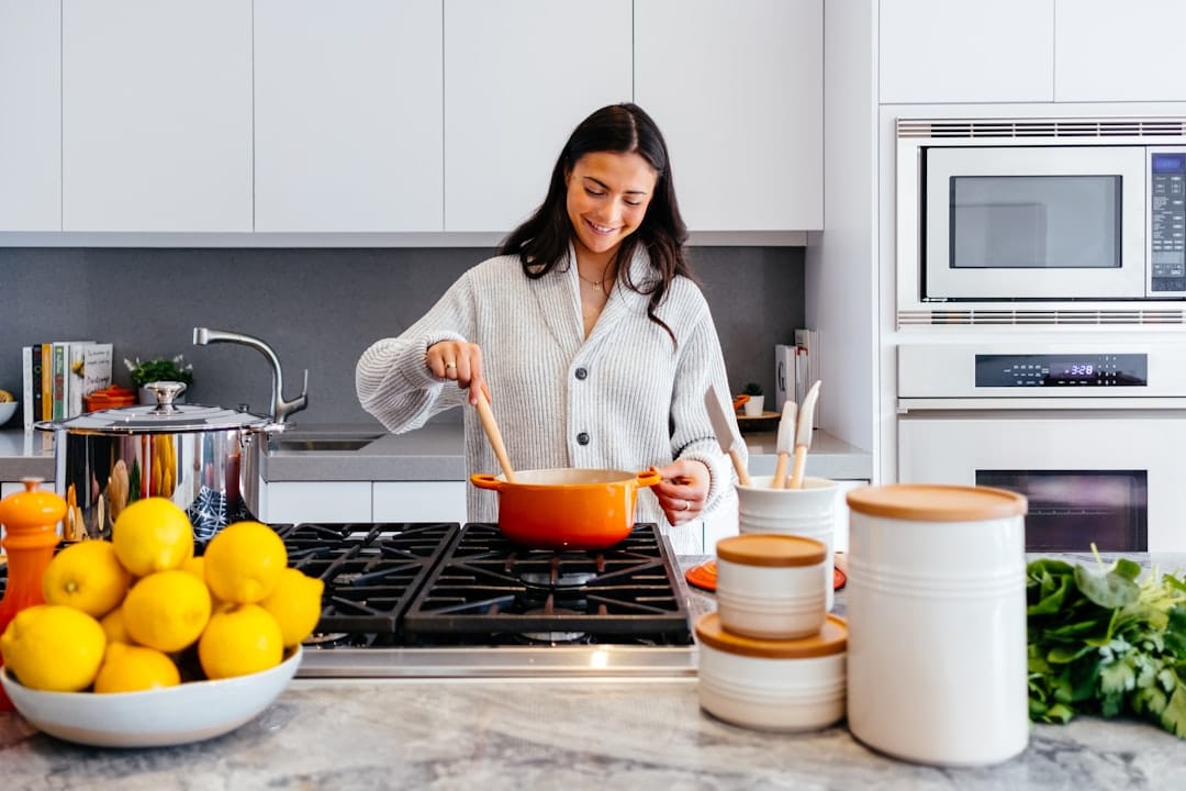 Smiling woman stirring an orange pot on a gas stove in a bright kitchen with a bowl of lemons