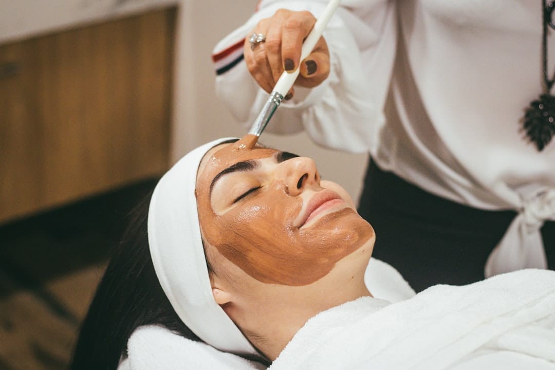Woman reclining in a salon while a therapist brushes brown clay onto her face