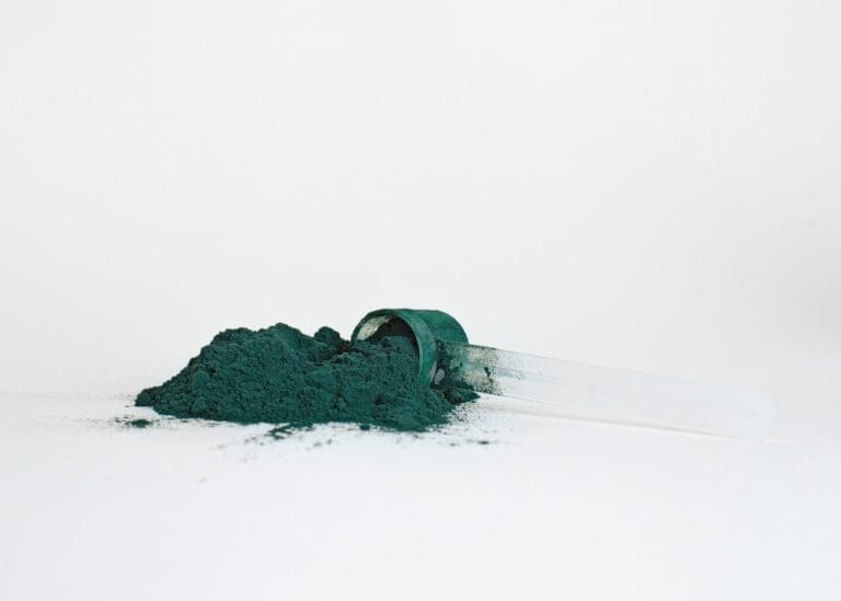 Dark blue-green algae powder in a heap beside a clear plastic measuring scoop on a white surface