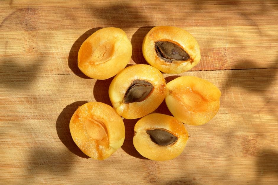 Six halved fresh apricots on a wooden board, three still holding their brown stones