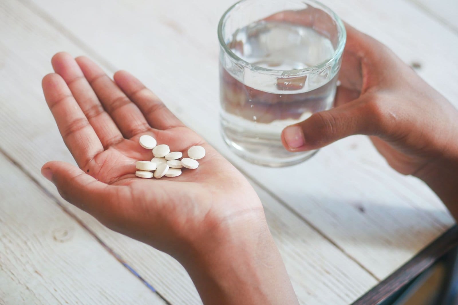 An open palm holding several small white tablets, with a glass of water in the other hand
