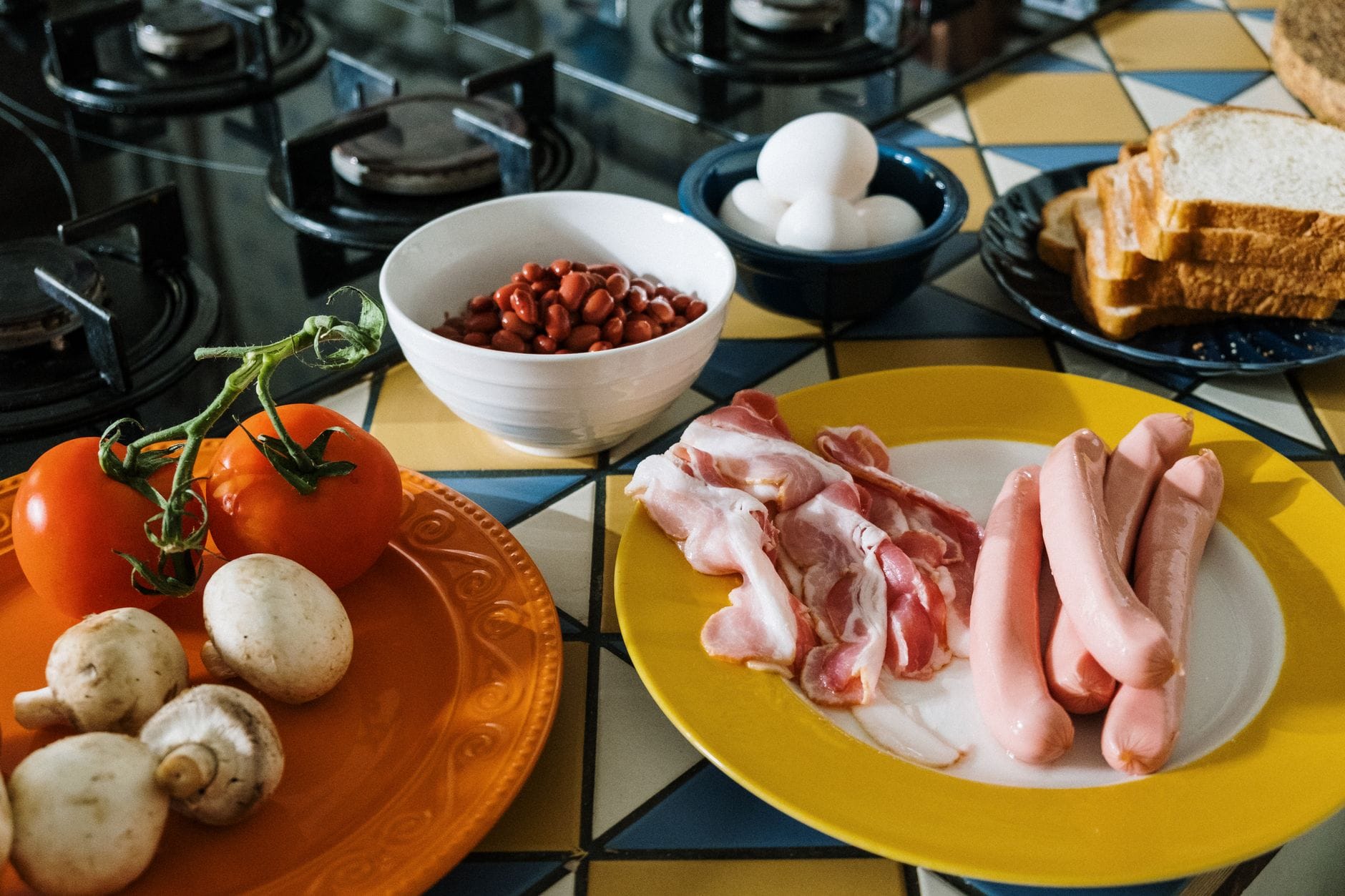 Uncooked bacon rashers and frankfurter sausages on a yellow plate, with tomatoes, mushrooms, beans, eggs and sliced bread