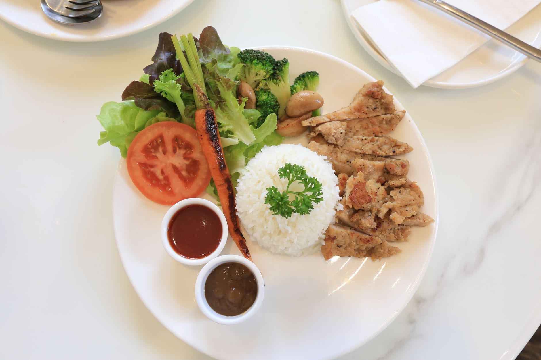 White plate with sliced grilled meat, white rice, salad leaves, broccoli, tomato and a roasted carrot