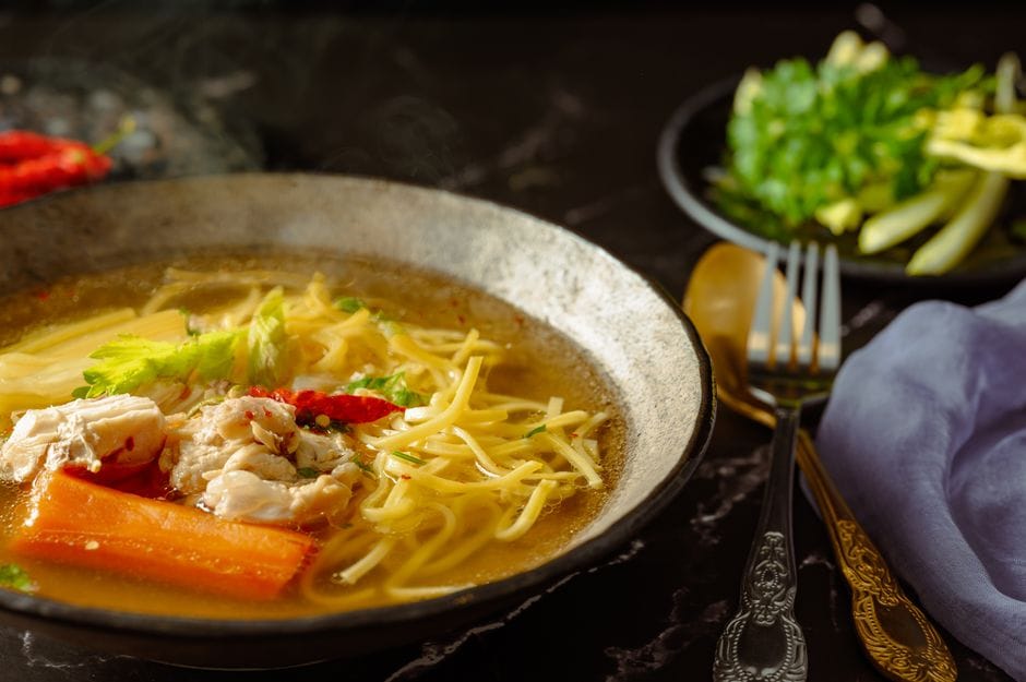 Bowl of steaming chicken noodle soup with shredded chicken, carrot, celery and noodles beside gold cutlery