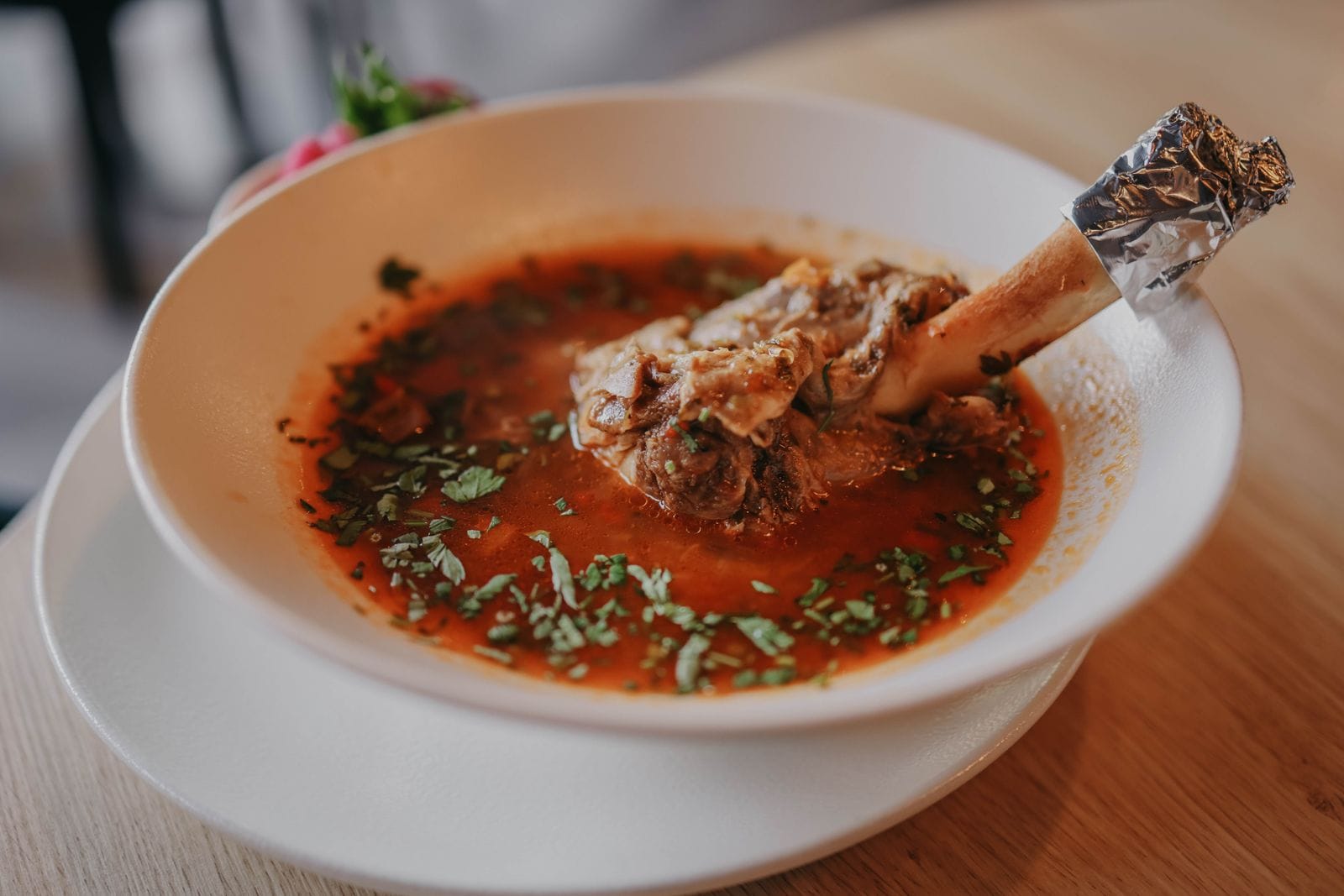 A lamb shank standing in a bowl of clear red broth scattered with chopped herbs, the bone and connective tissue visible