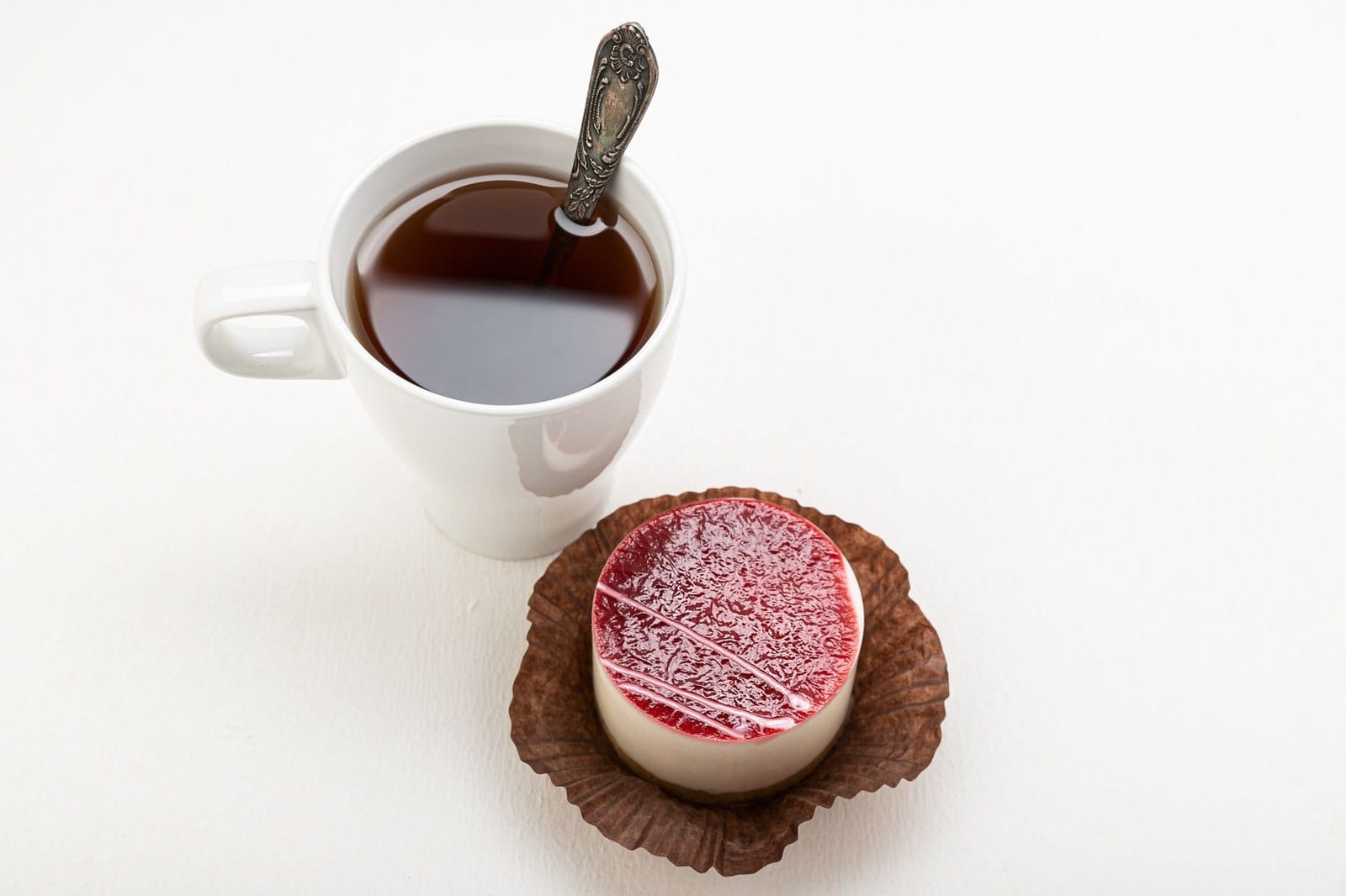 A small round dessert topped with a red fruit jelly glaze, served beside a cup of tea
