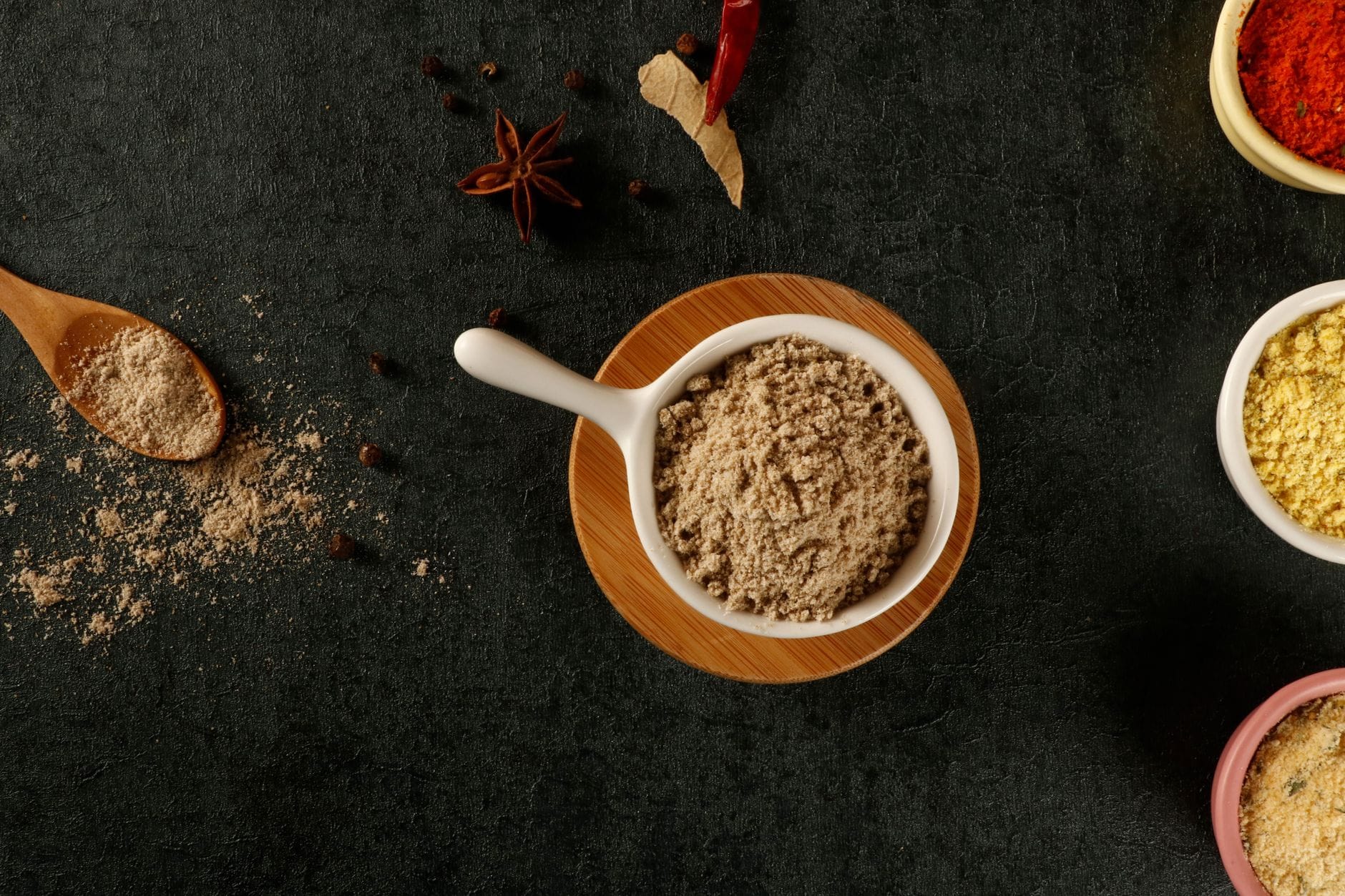 Pale ground spice powder in a small white dish on a dark surface, with star anise and dried chilies nearby