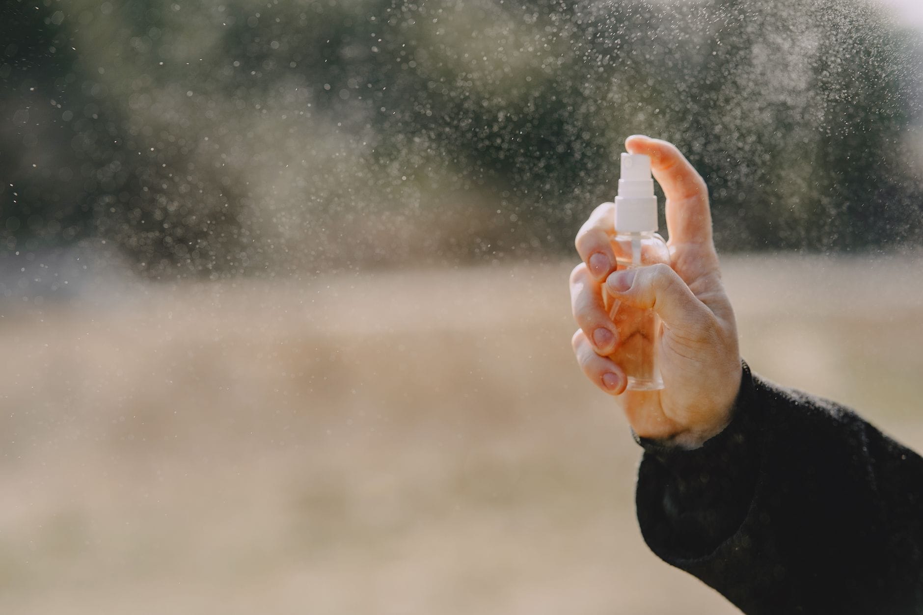 Hand spraying a small bottle outdoors, with mist catching the light