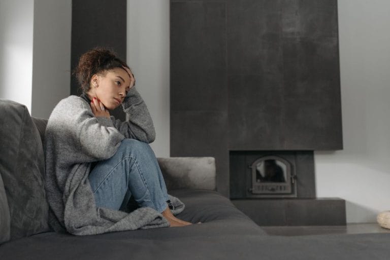 Woman sitting curled up on a grey sofa in a grey cardigan, one hand at her head and one at her throat, looking unwell