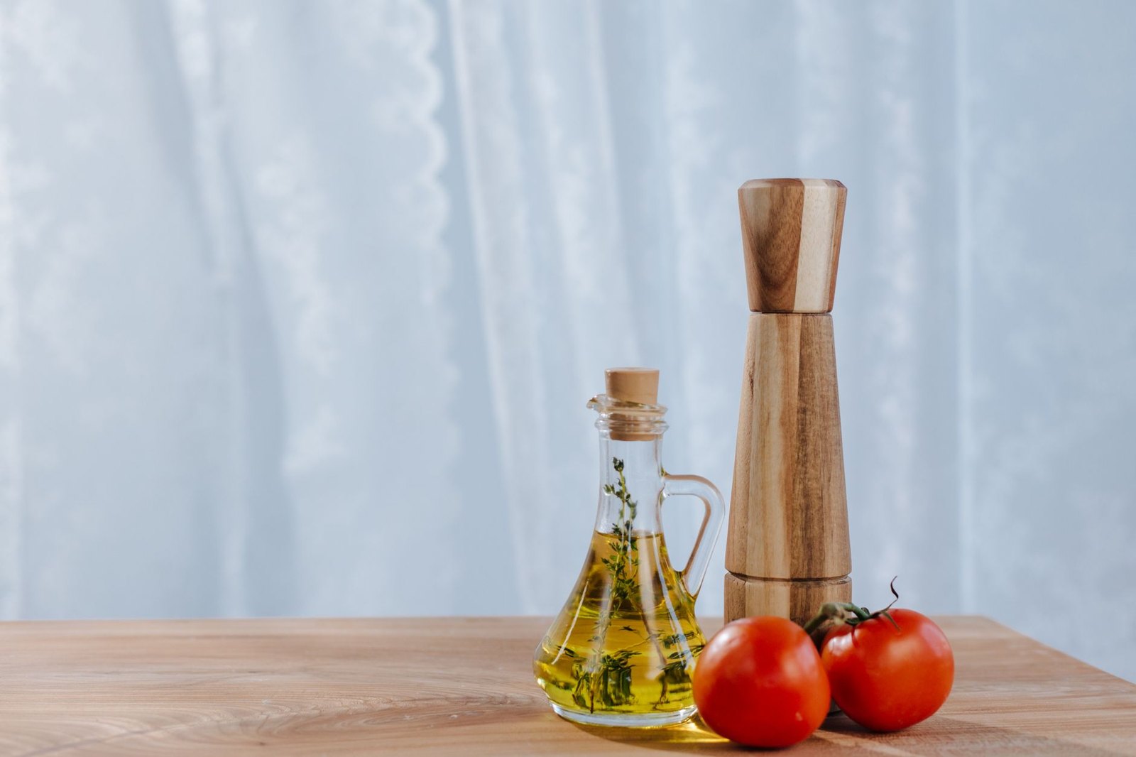 A glass cruet of herb-infused oil beside a wooden pepper mill and two tomatoes on a table