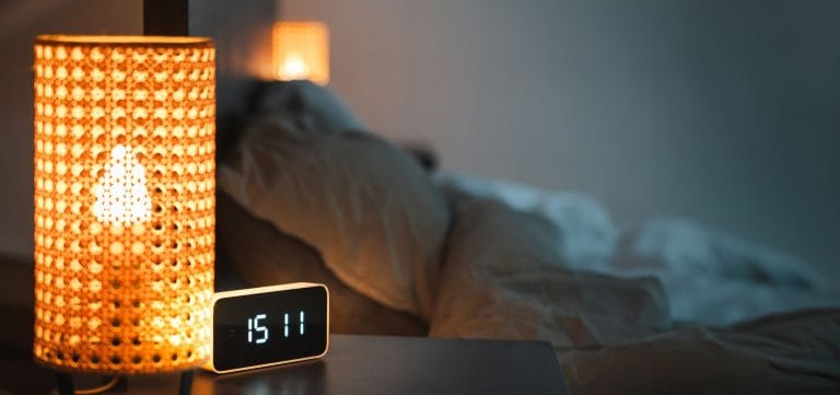 Bedside lamp glowing beside a digital alarm clock with an unmade bed behind