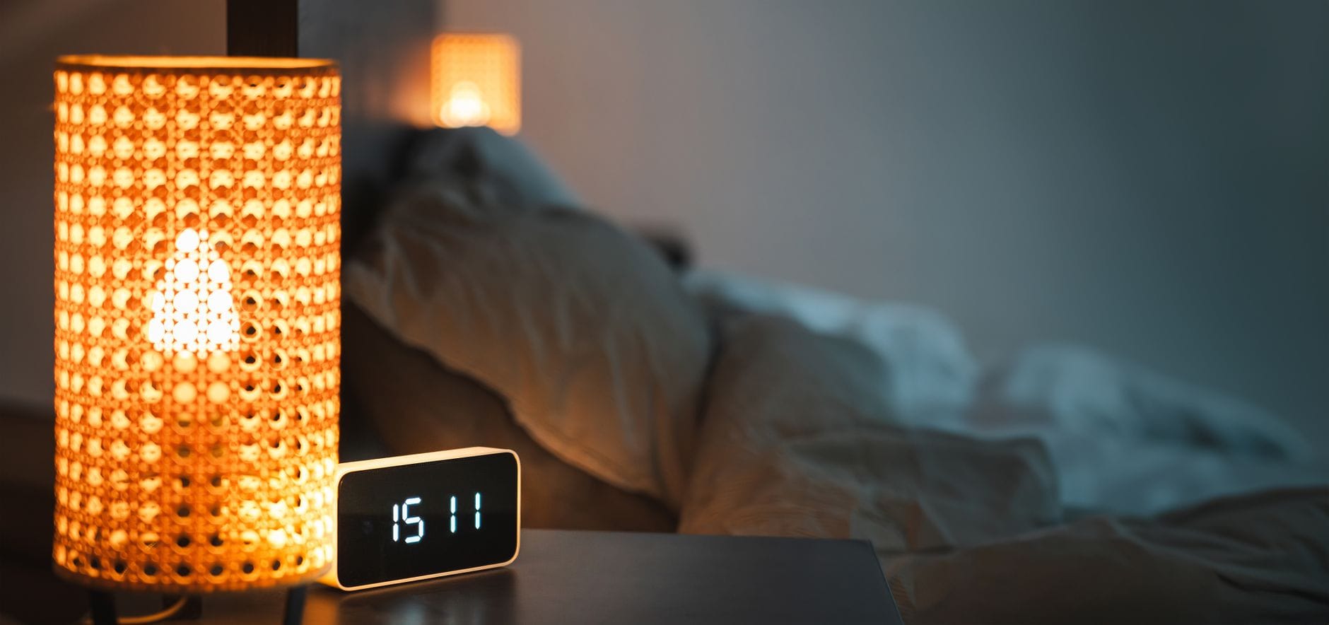 Bedside lamp glowing beside a digital alarm clock with an unmade bed behind