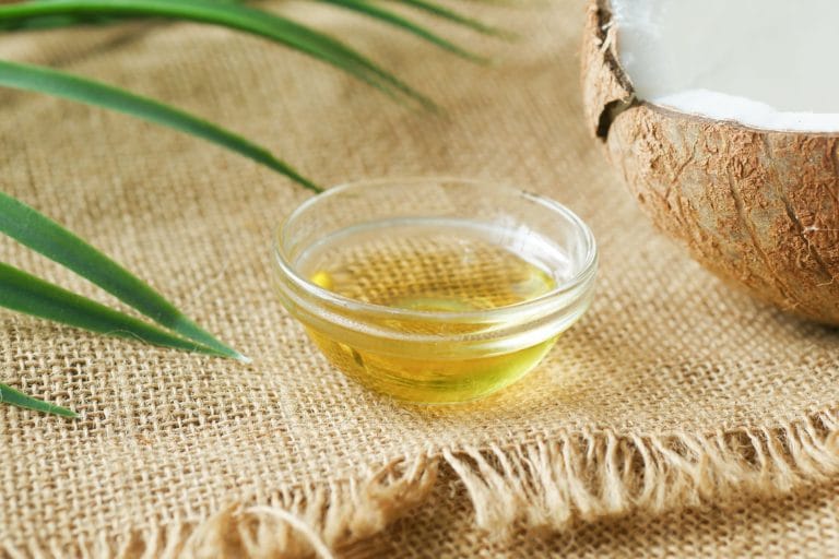 Small glass bowl of clear liquid oil on burlap beside a halved coconut and palm leaf