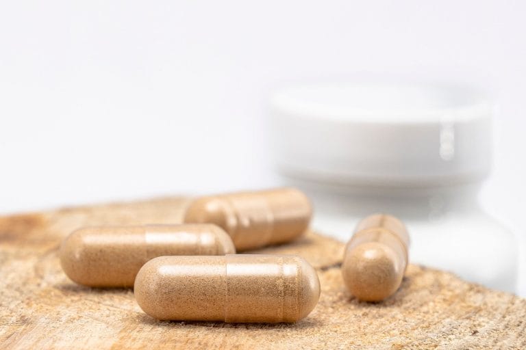 Four brown herbal supplement capsules on a wooden surface with a white bottle blurred behind