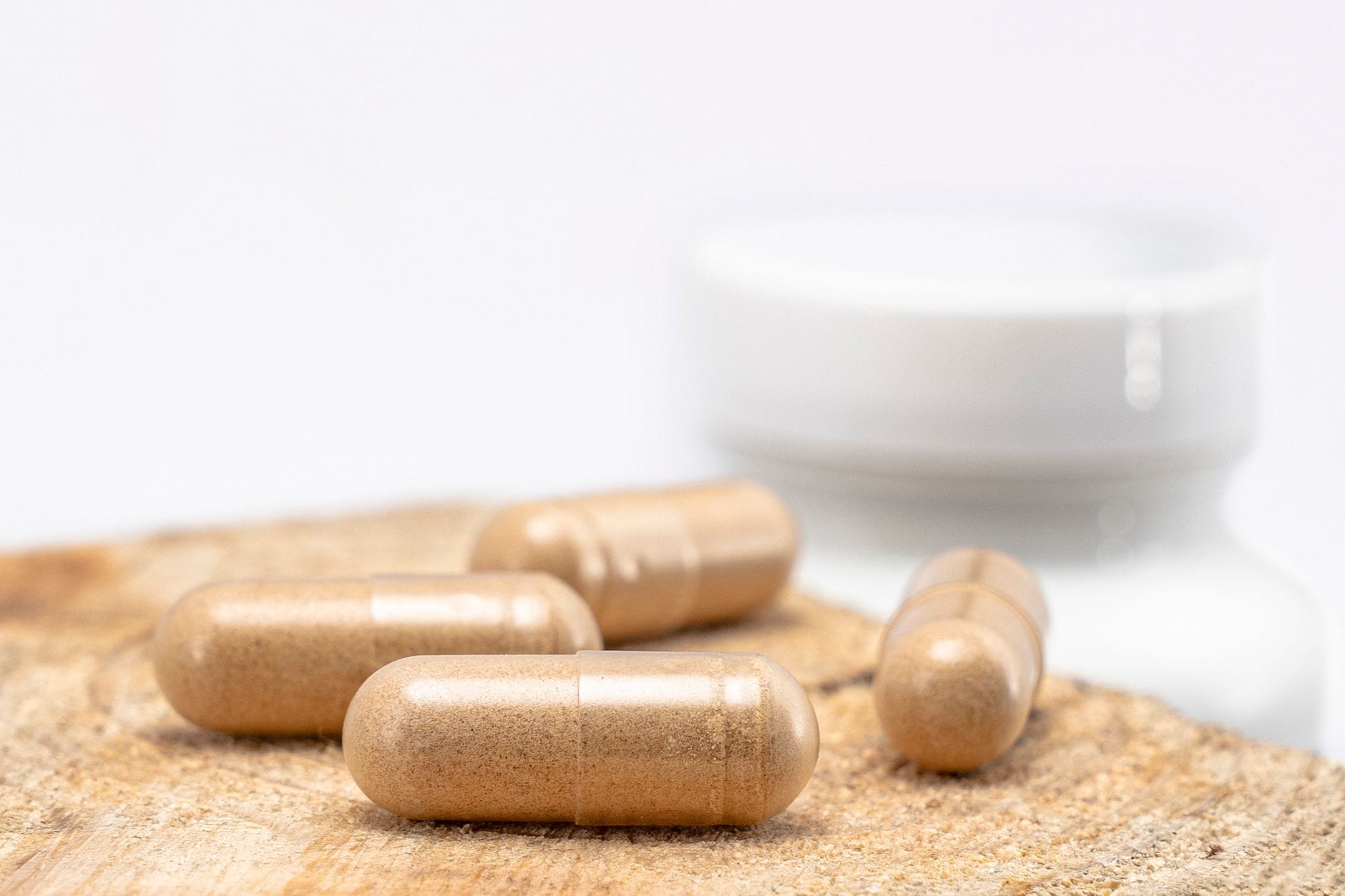 Four brown herbal supplement capsules on a wooden surface with a white bottle blurred behind