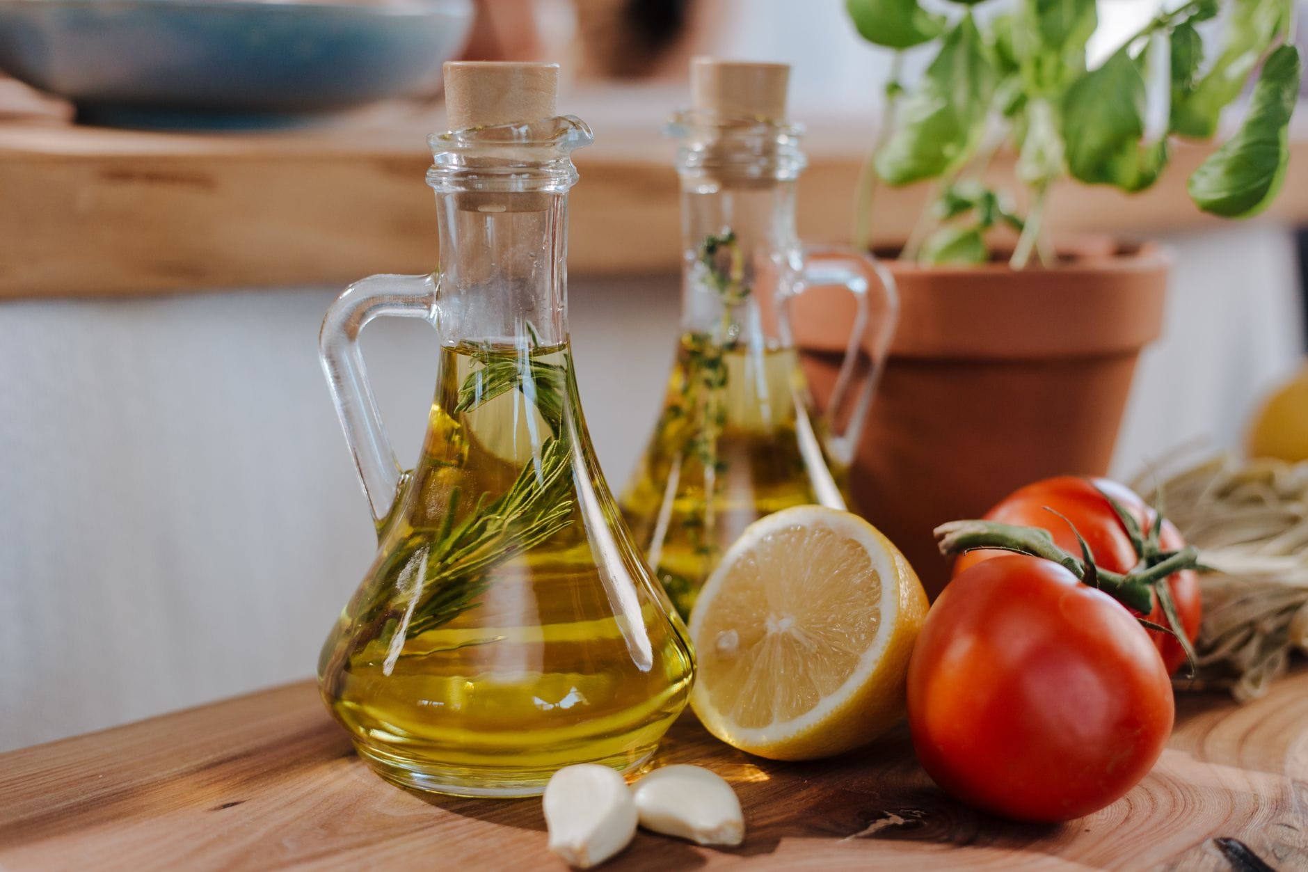 Glass cruets of herb-infused olive oil on a wooden board with a lemon, tomatoes and garlic