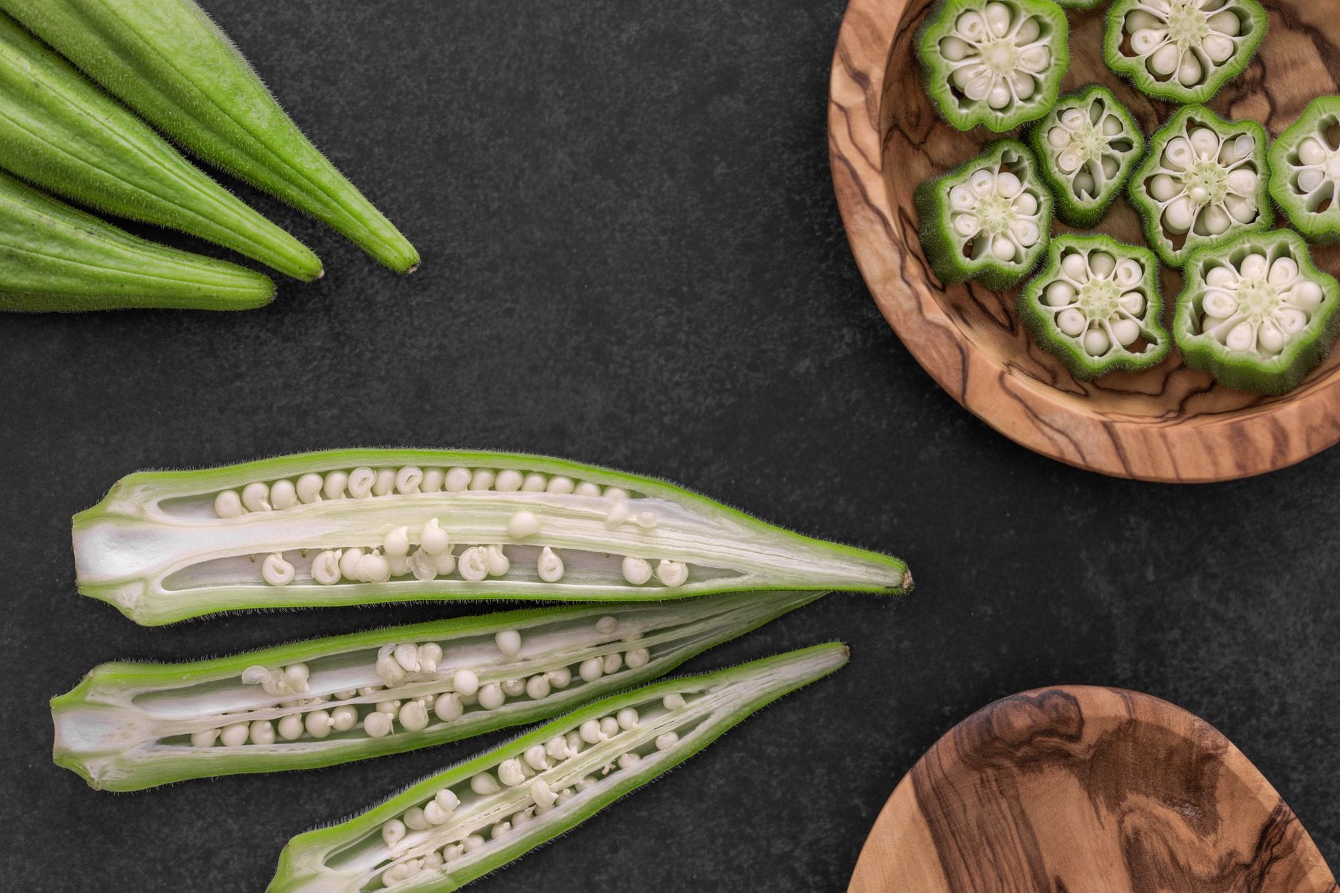 Whole, halved and sliced green okra pods with white seeds on slate