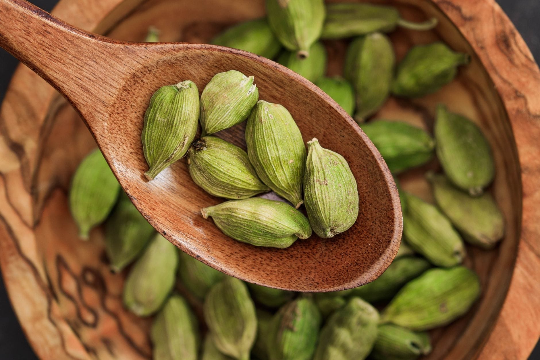 Whole green cardamom pods on a wooden spoon above a wooden bowl