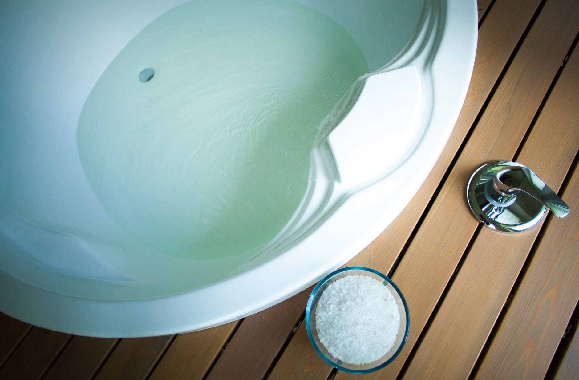 Bathtub filled with warm water beside a glass bowl of coarse bath salt