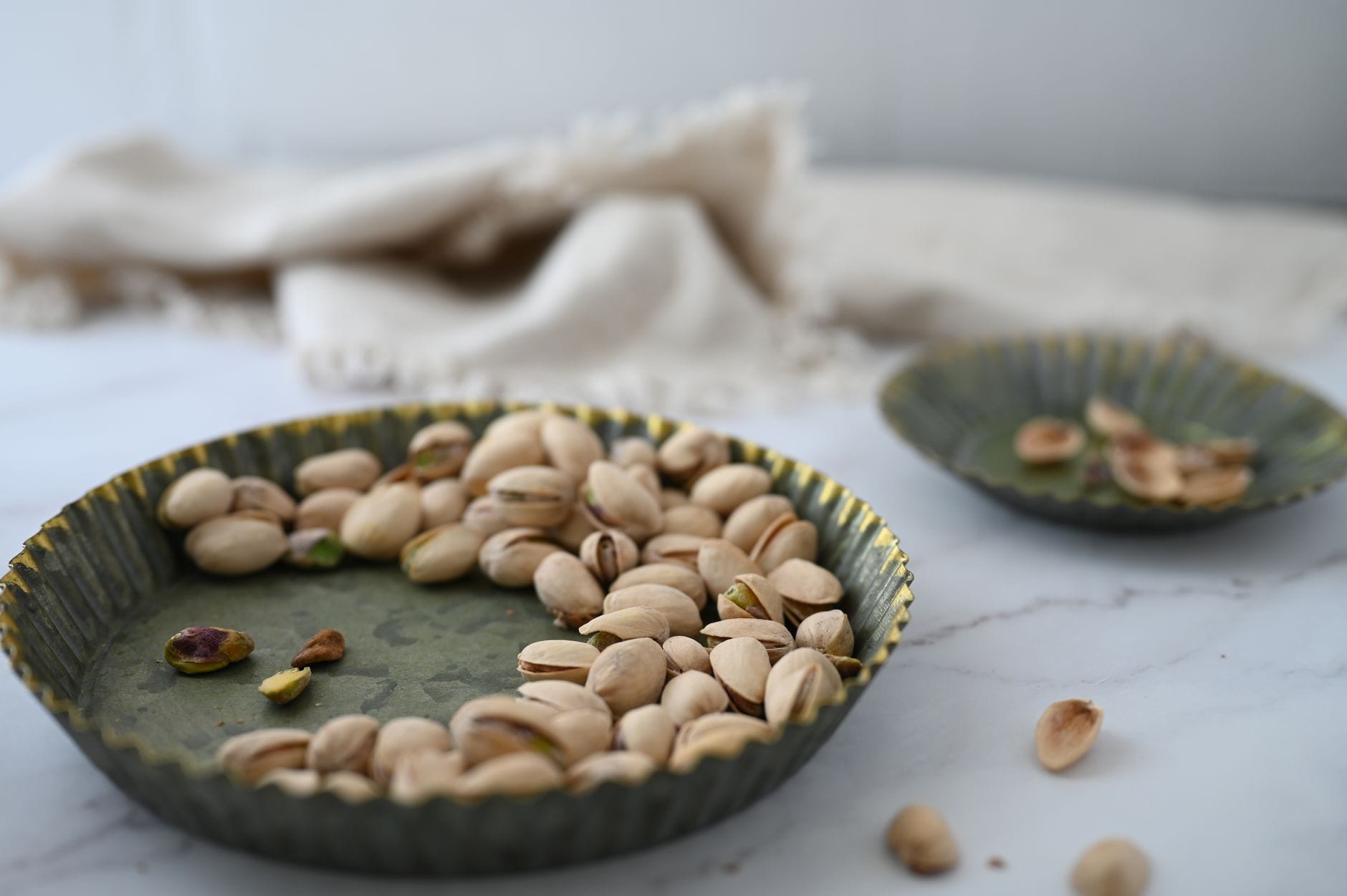 In-shell pistachios in a green fluted metal dish on marble with empty shells in a second dish
