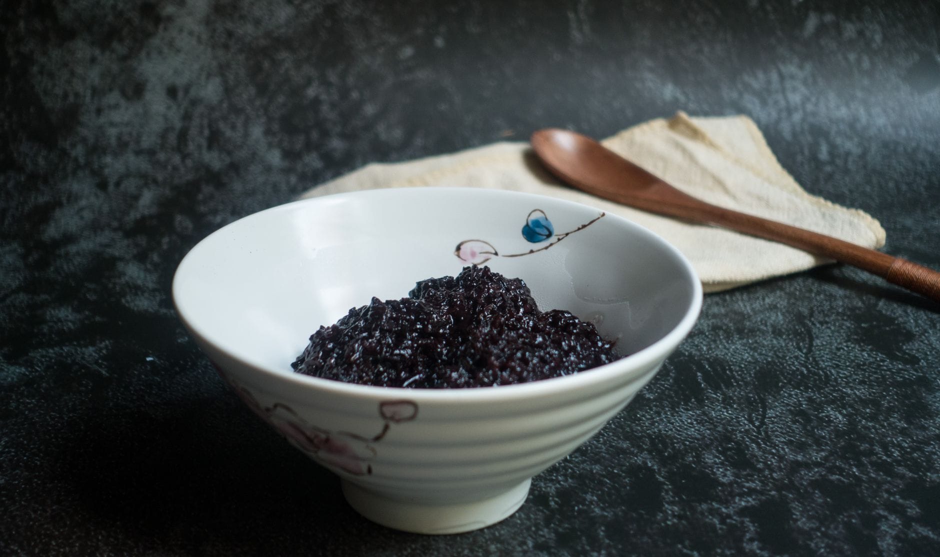 White ceramic bowl of cooked black rice with a wooden spoon on a dark stone surface