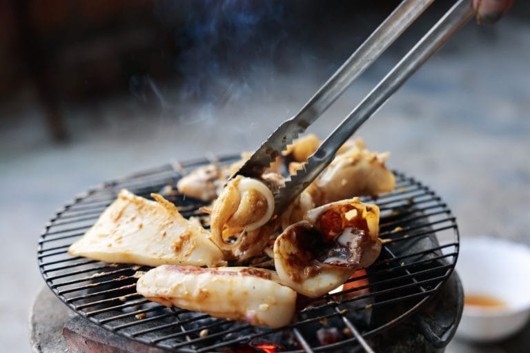 Squid pieces and tentacles cooking on a round charcoal grill, turned with metal tongs