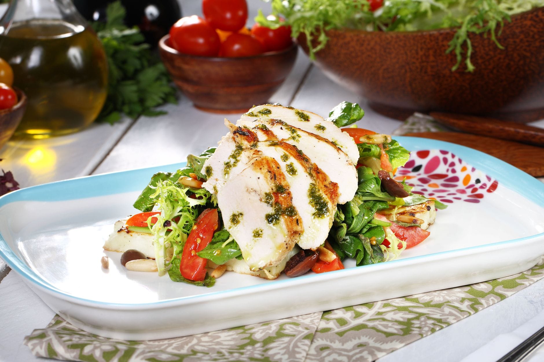 Sliced grilled chicken breast with herb sauce on a leafy green salad with tomatoes, olives and pine nuts, olive oil bottle behind
