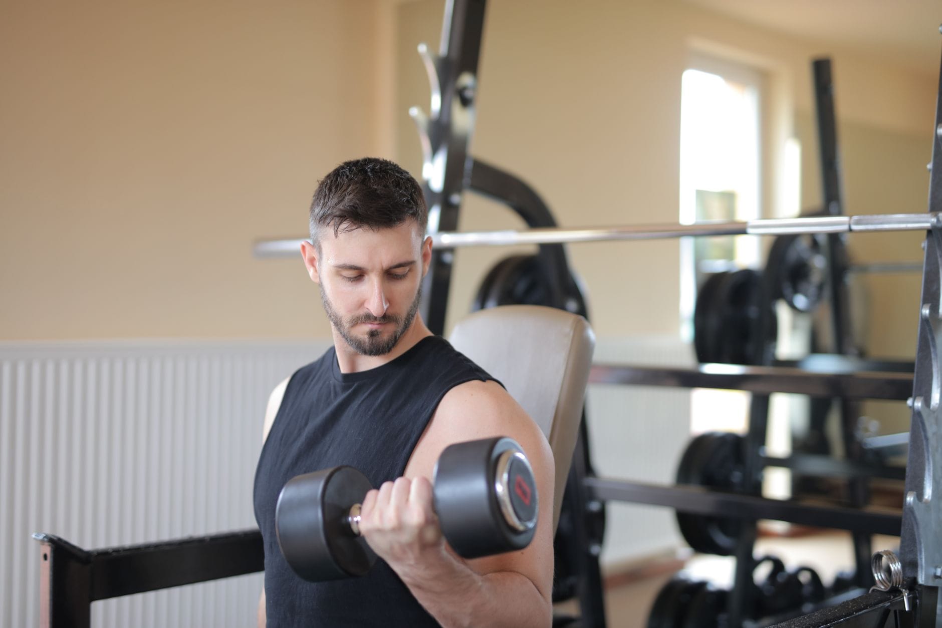 Man curling a black dumbbell in a plain gym beside a weight rack
