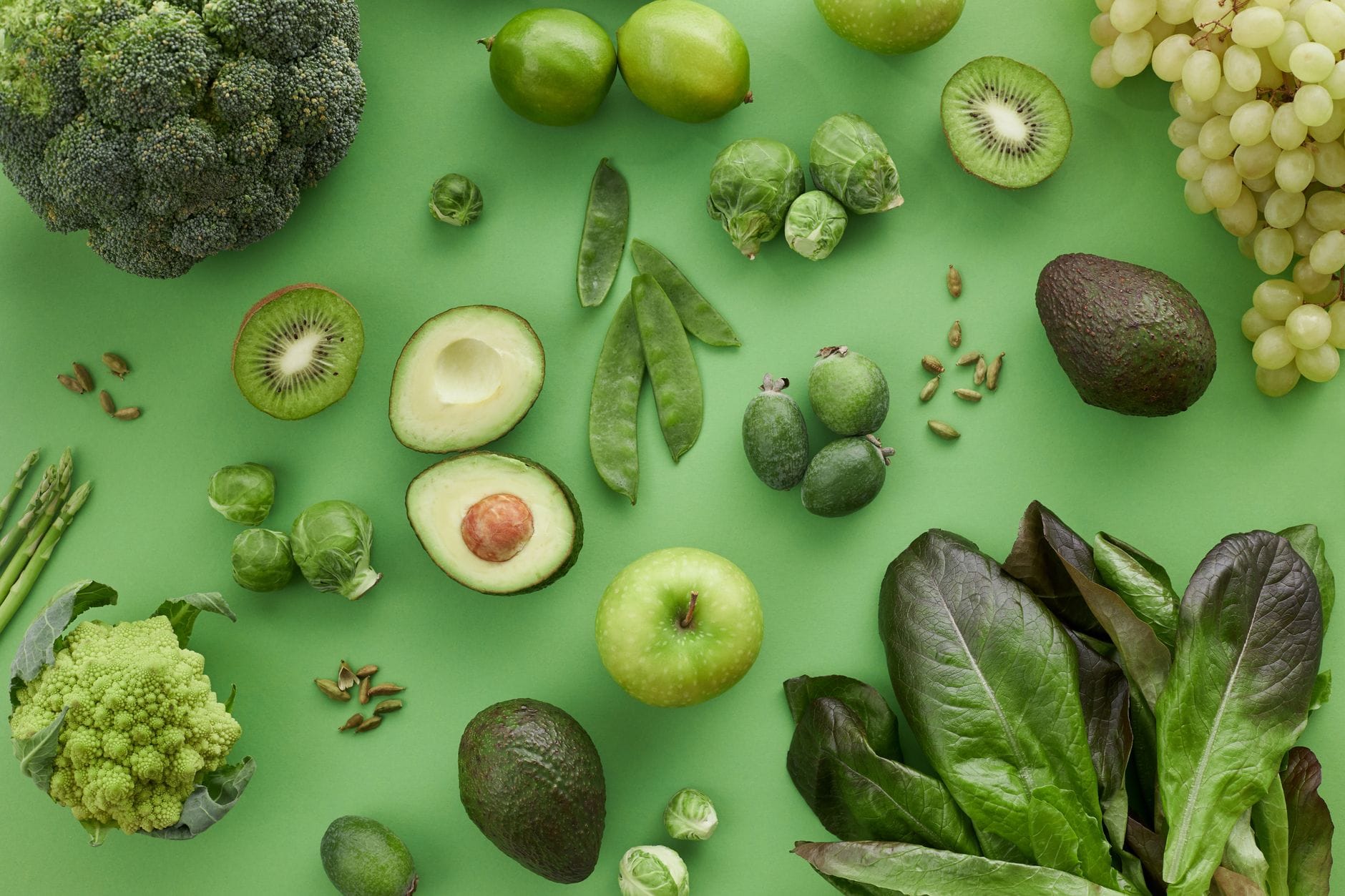 Flat lay of green produce on a green background including broccoli, avocado, kiwi, green apple, grapes and leafy greens