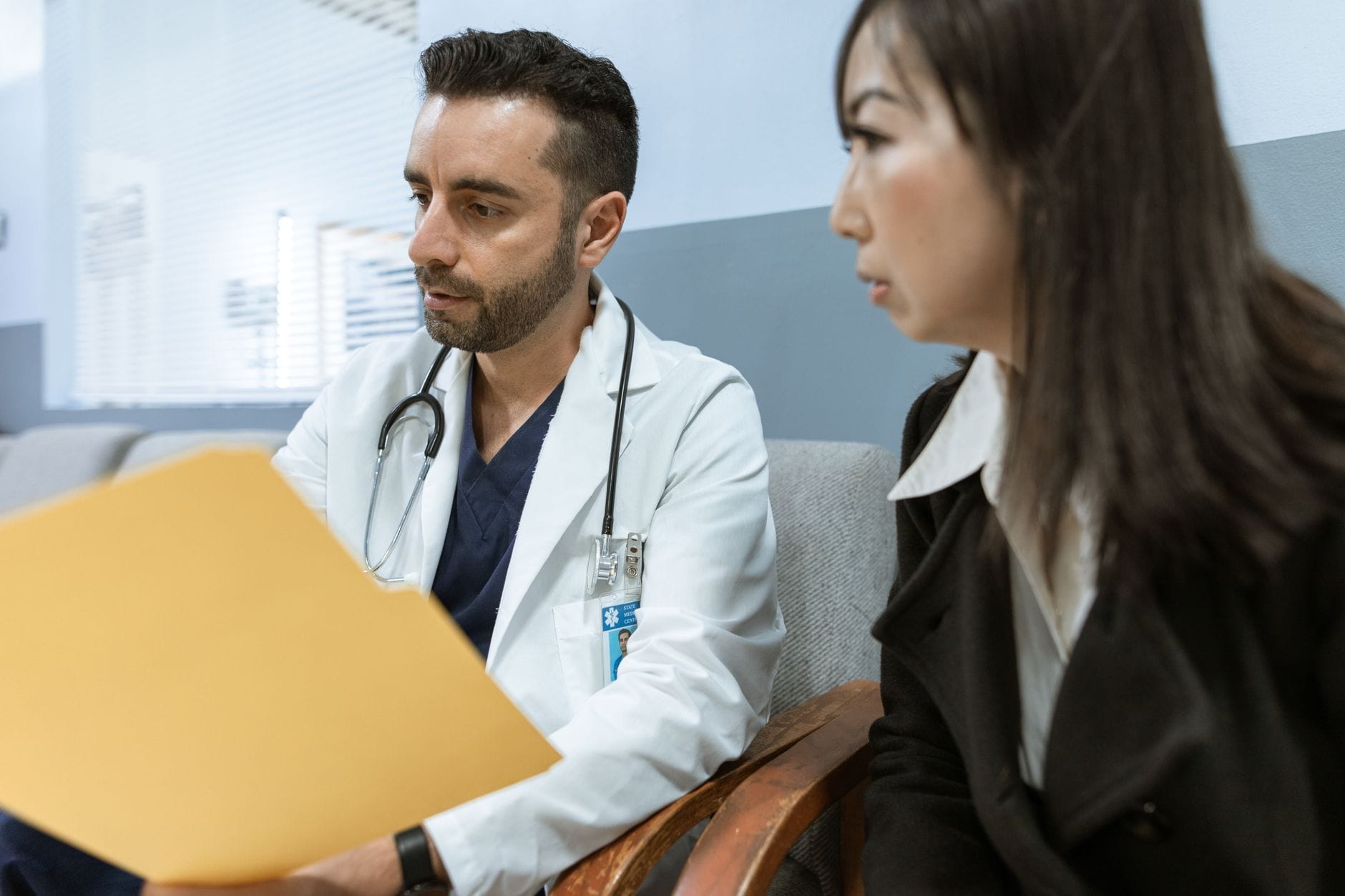 Doctor in a white coat with a stethoscope reviewing a folder beside a seated patient in a clinic