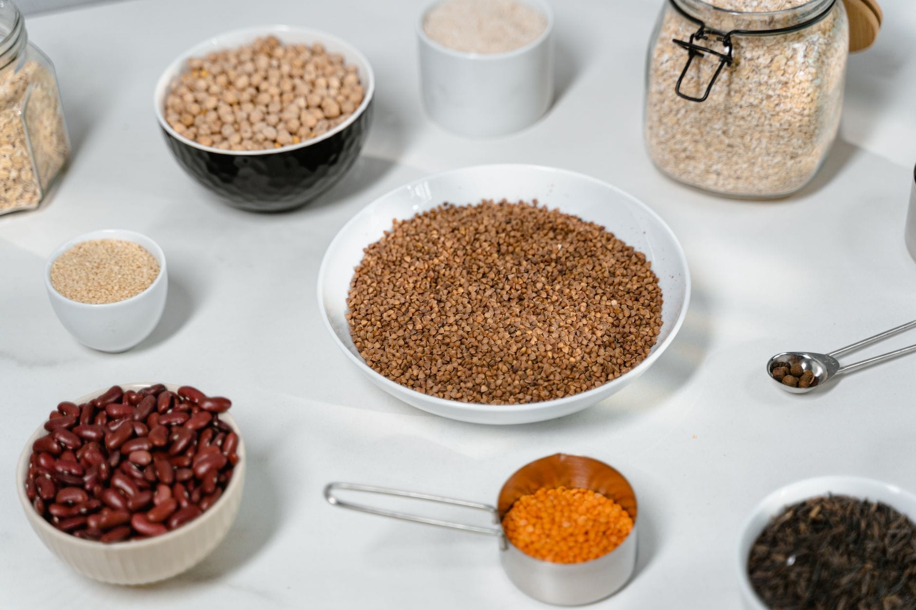 Bowls of chickpeas, kidney beans, buckwheat, red lentils and oats on a white surface