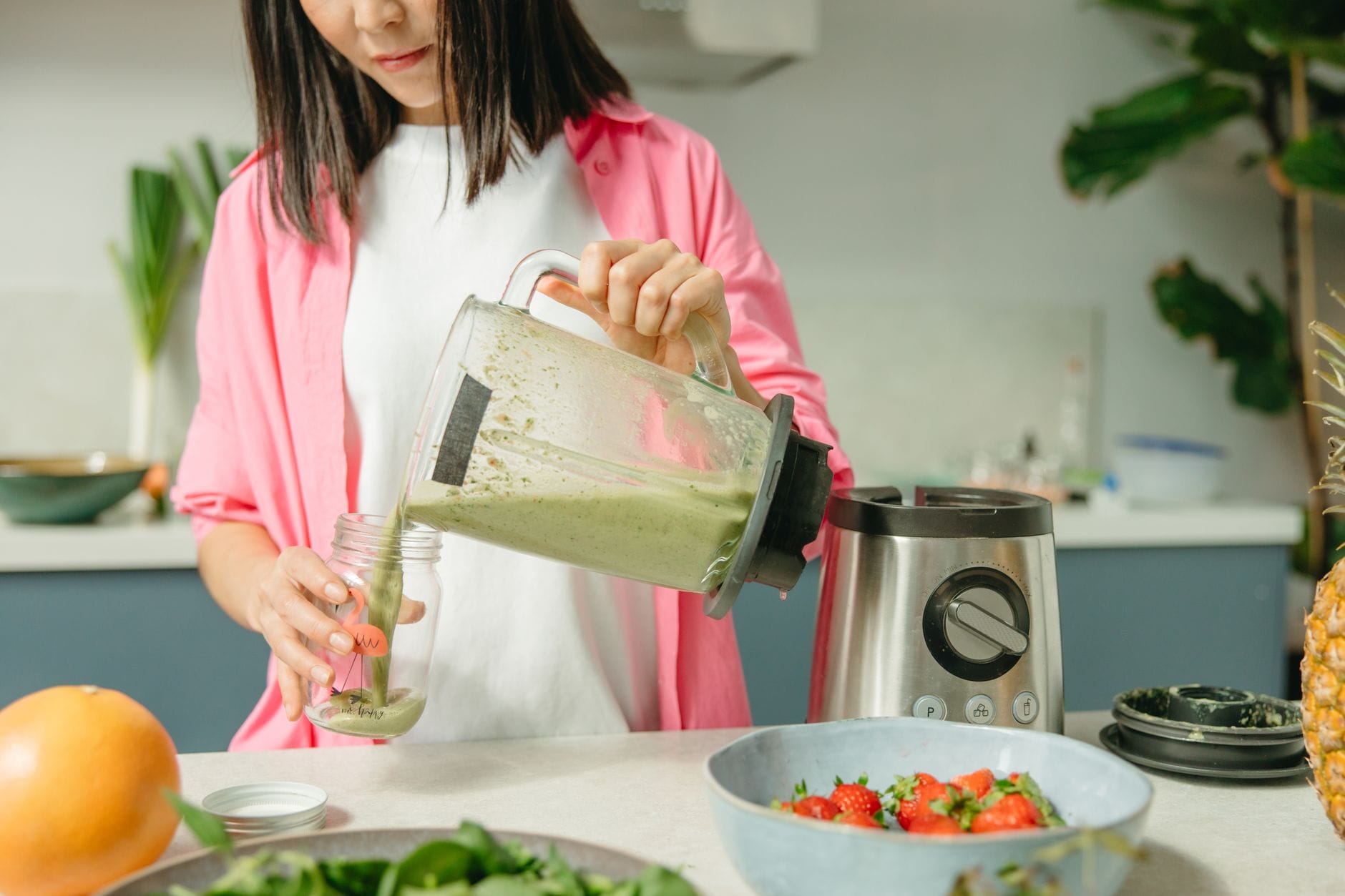 Woman pouring a green smoothie from a blender jug into a glass jar in a kitchen