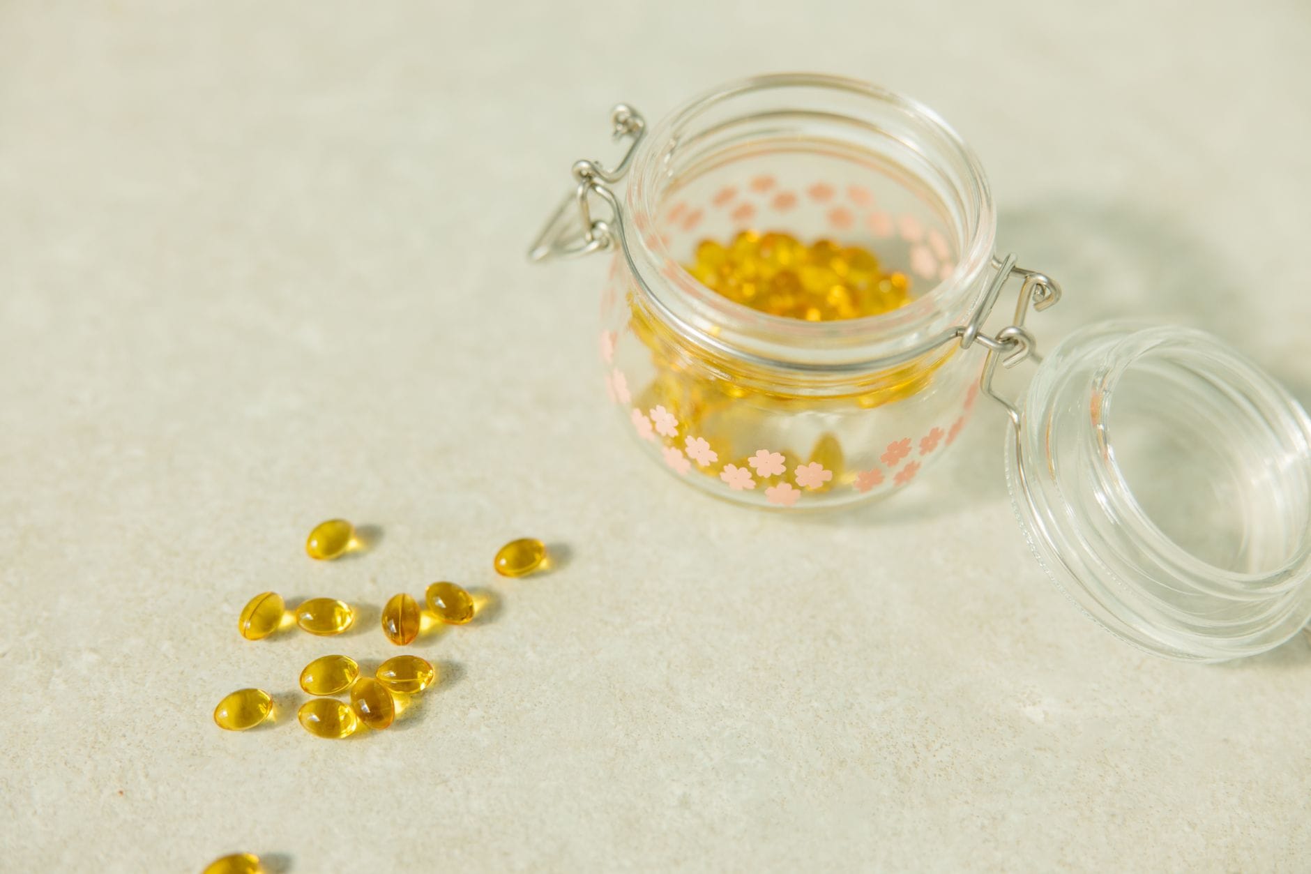 Open glass jar of yellow softgel capsules with several spilled on a pale surface