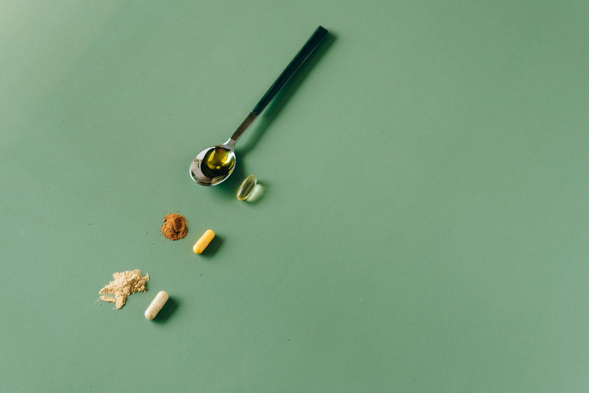 Spoon of oil capsule beside two small piles of supplement powder and capsules on a green background