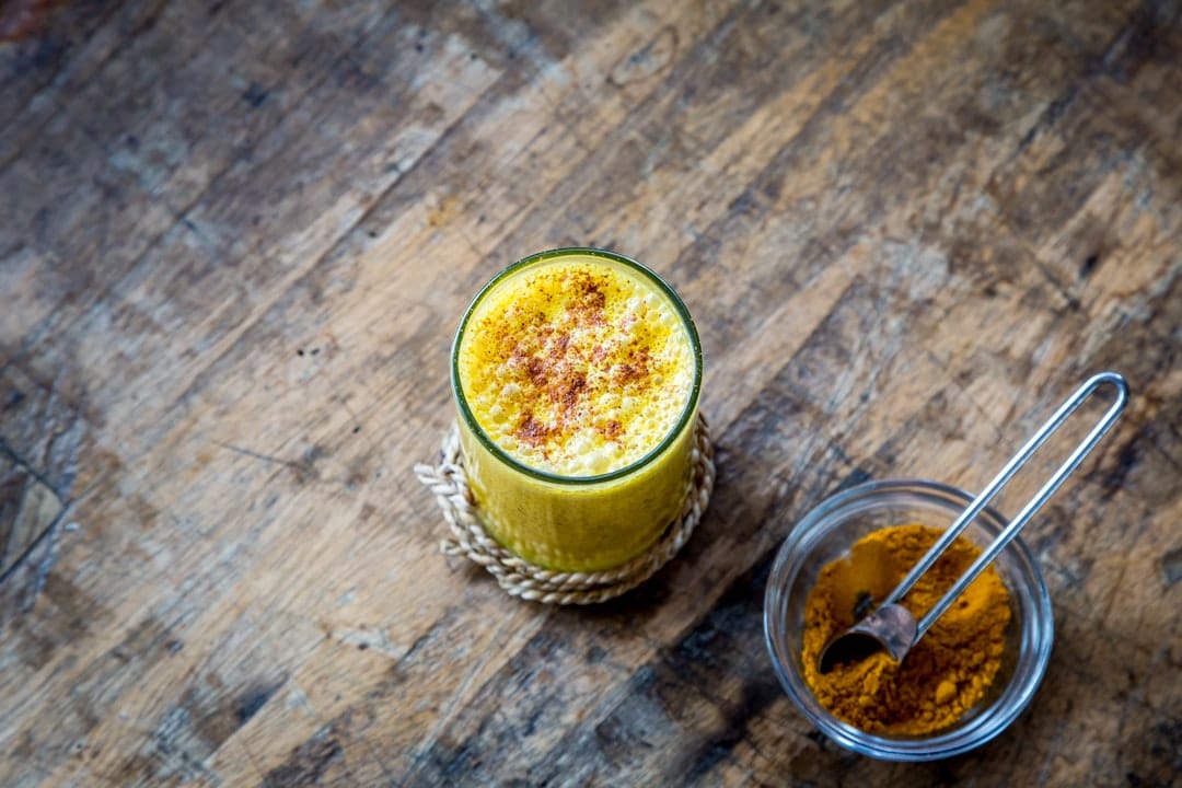 Glass of frothy golden turmeric milk dusted with cinnamon on a wooden table, beside a small glass bowl of ground turmeric with a spoon