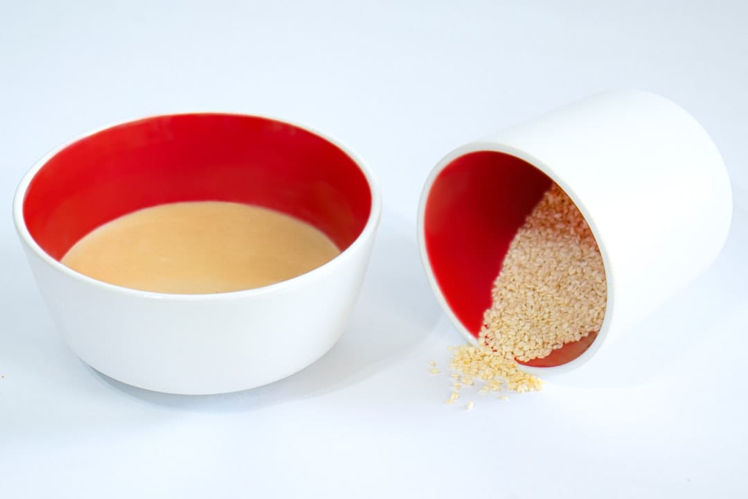 Bowl of smooth pale tahini beside a tipped bowl spilling sesame seeds